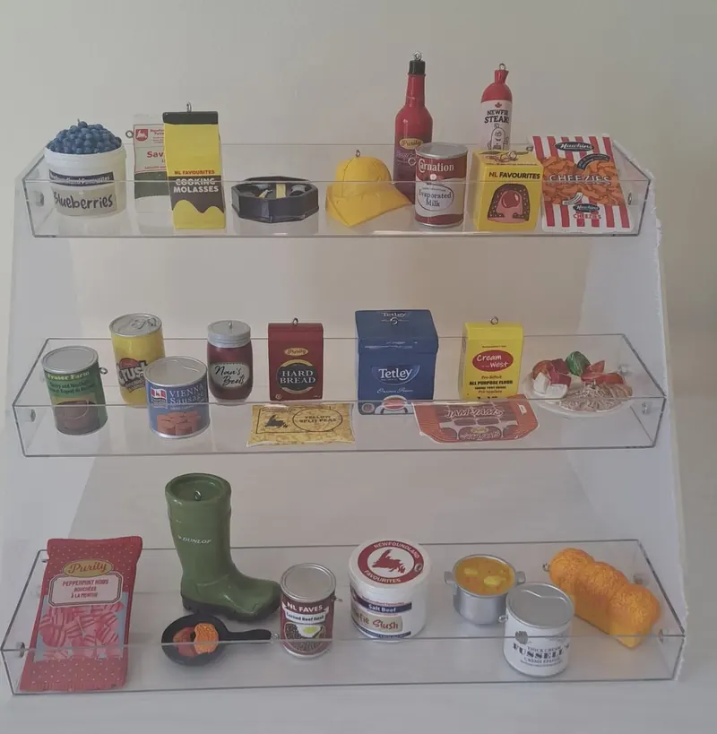 Three clear shelves of miniature Newfoundland ornaments: a green Dunlop boot, Tetley tea, cooking molasses, tins, a loaf of bread and more